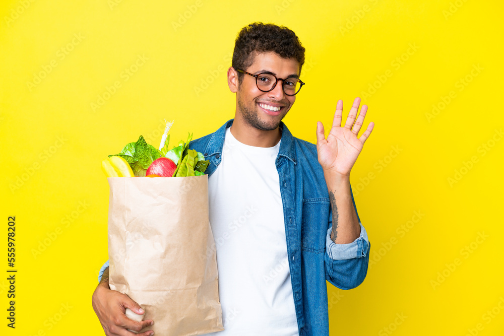 Young Brazilian man holding a grocery shopping bag isolated on yellow background saluting with hand with happy expression Young Brazilian man holding a grocery shopping bag isolated on yellow background saluting with hand with happy expression