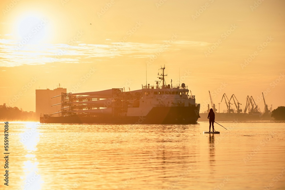 Girl standing on paddle board near cargo vessel loaded with rotor blades for wind turbines at the sunrise Danube river Girl standing on paddle board near cargo vessel loaded with rotor blades for wind turbines at the sunrise Danube river