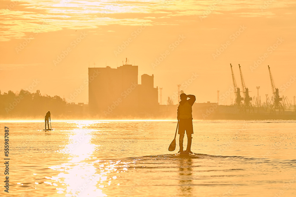 Orange winter sunset and silhouette of boy rowing on supboards (SUP) at calm Danube river at the morning against port cranes