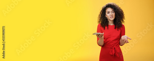 So waht, I confused. Questioned uncertain woman with curly hair in red dress shrugging looking clueless as holding hand cannot understand where order as checking mail box via device over yellow wall