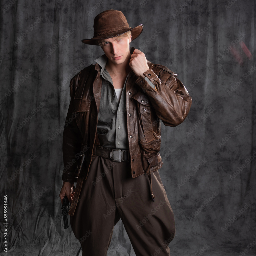 A young man in retro style, an adventure character. The hero of the adventure, a guy in a hat and a leather jacket, posing in the studio on a gray background - obrazy, fototapety, plakaty 