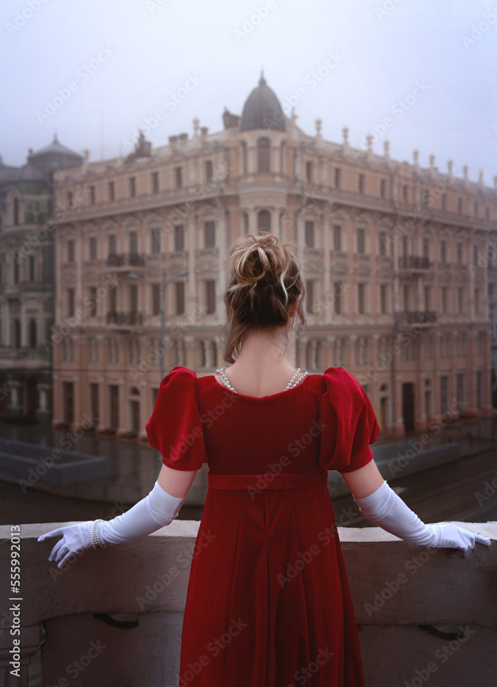 Beautiful young girl in a nineteenth century dress stands on a balcony in front of a beautiful building.