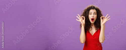 Woman assring husband he doing great supporting him during poket match standing amazed and excited over purple wall in attractive red dress showing okay or excellent sign folding lips from excitement photo