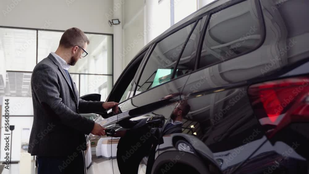 A stylish man in glasses and suit closes the door of the new car in car dealership. A man examine vehicle before making purchase. Buy car A stylish man in glasses and suit closes the door of the new car in car dealership. A man examine vehicle before making purchase. Buy car