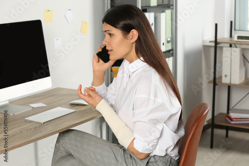 Young woman with arm wrapped in medical bandage talking on phone at workplace