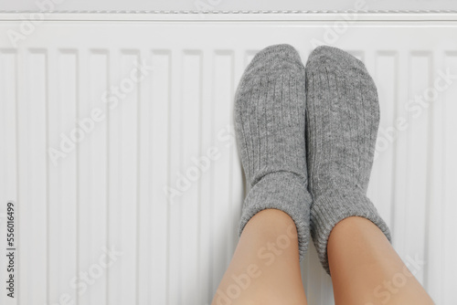Woman warming legs on heating radiator near white wall, closeup. Space for text