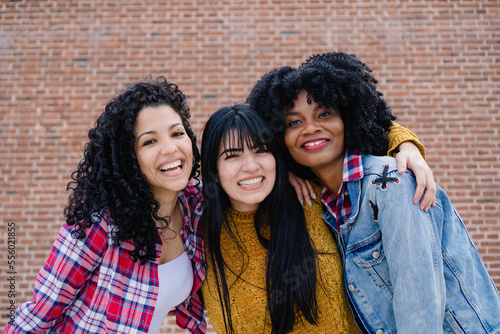 Three young, Latina, Hispanic, multiracial friends smiling and hugging while posing for a portrait. Brick background
