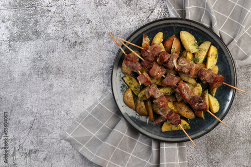 Kebabs on wooden skewers with potatoes and greens on a round plate on a dark gray background. Top view, flat lay