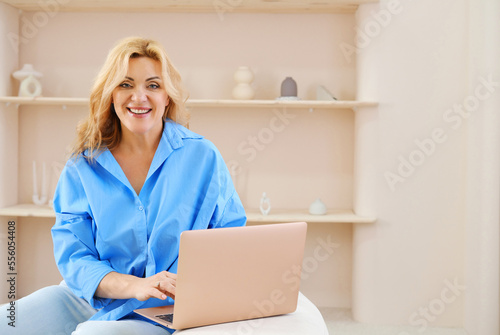 Beautiful mature blonde woman in a blue shirt sits on a sofa with a laptop in a beige color interior, looks at the camera and smiles