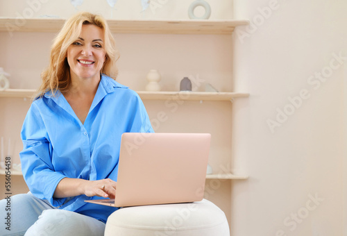 Beautiful mature blonde woman in a blue shirt sits on a sofa with a laptop in a beige color interior, looks at the camera and smiles