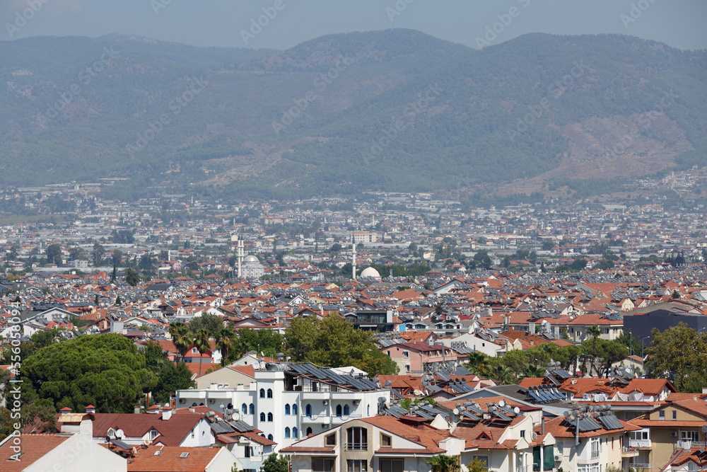 A view of the city from above against a background of towering mountains and hills. Panorama of the Turkish city of Fethiye with mosques and minarets. Tiled roofs of houses with solar panels