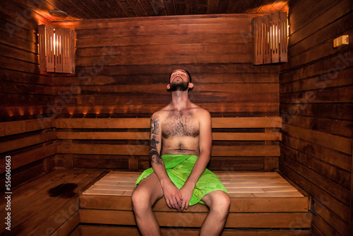 Young bearded man in green towel sitting on bench in bathhouse sauna and relaxing, gets high. Guy resting in finnish sauna at wooden wall. Wellness, self care, healthy concept. Copy text space