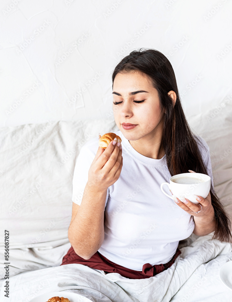 Young brunette woman sitting in the bed with eating croissants Young brunette woman sitting in the bed with eating croissants