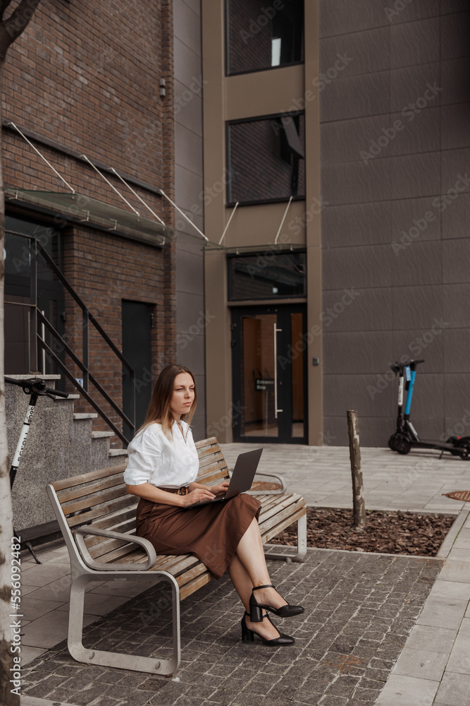 Young business woman sitting on a bench with a laptop of the modern office building background Young business woman sitting on a bench with a laptop of the modern office building background