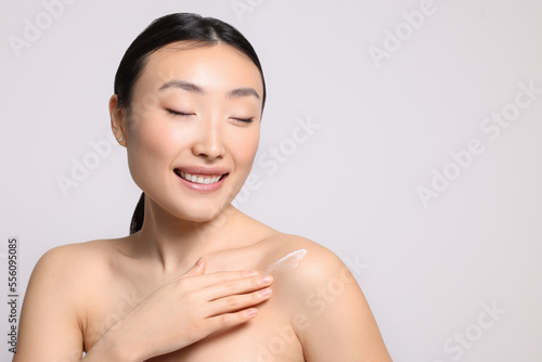 Beautiful young Asian woman applying body cream onto shoulder on light grey background, space for text
