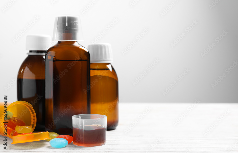 Bottles of syrup, measuring cup, lozenges and spoon on white wooden table against light grey background, space for text. Cold medicine Bottles of syrup, measuring cup, lozenges and spoon on white wooden table against light grey background, space for text. Cold medicine