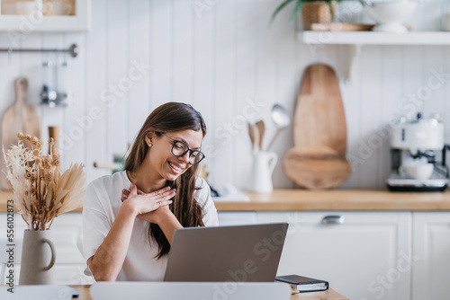 Картината върху платно Adorable female student in white t-shirt sitting at kitchen table with laptop puts hands on chest feels gratitude after sistant lesson