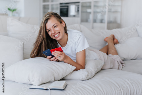 Cheerful European young adult businesswoman laying on sofa at home in casual holds phone and credit card makes purchase via internet looks at camera smiles wide. Successful people. Shopping online.