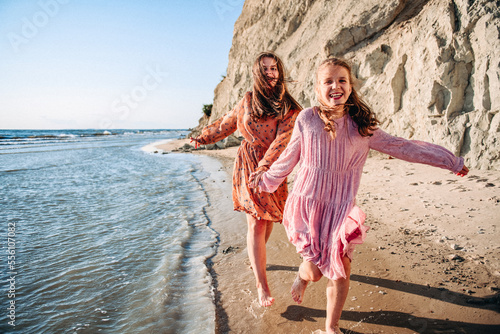 Billede på lærred Two happy smiling teen caucasian barefooted girls wearing airy dresses holding their hands and running along sandy coast with white rock in sunny summer day