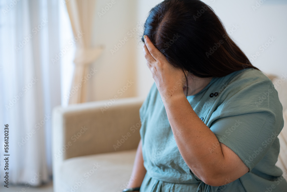 Indoor portrait with copy space of unrecognizable fat Asian woman sitting on sofa, holding temples, having headache. Migraine, shameless, sadness and depression concept. Indoor portrait with copy space of unrecognizable fat Asian woman sitting on sofa, holding temples, having headache. Migraine, shameless, sadness and depression concept.