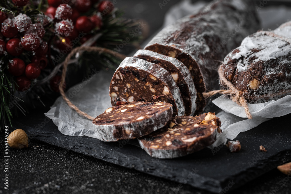 Chocolate salami filled with almonds, hazelnuts and raisins on the black background. Italian sweet sausage shaped dessert sprinkled with powdered sugar. Christmas and New Year festive pastry Chocolate salami filled with almonds, hazelnuts and raisins on the black background. Italian sweet sausage shaped dessert sprinkled with powdered sugar. Christmas and New Year festive pastry