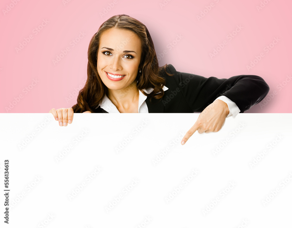Cheerfully smiling brunette business woman standing behind, peeping from blank banner or mock up signboard, showing copy space for text, pink color background. Cheerfully smiling brunette business woman standing behind, peeping from blank banner or mock up signboard, showing copy space for text, pink color background.
