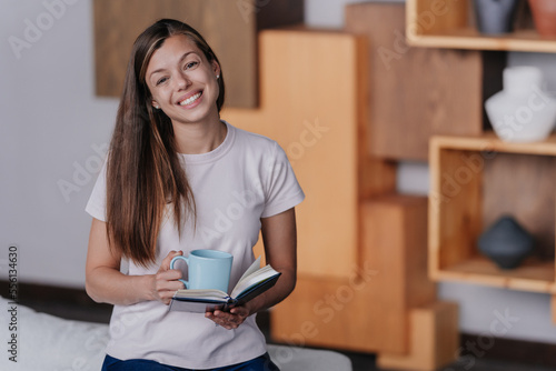 Beautiful Italian student girl in white t-shirt holds notebook drinks coffee at home. Happy hispanic young woman at morning prepares for work. Satisfied female with cup of coffee. Domestic leisure.