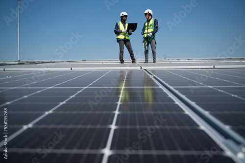 Caucasian and African American engineer man use electric screwdriver and computer notebook working with solar panel on roof factory