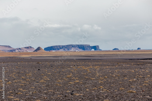 Dyrhólaey visto dall'entroterra, il suo arco si staglia sulla distesa di sabbia da cui escono ciuffi d'erba rossa photo