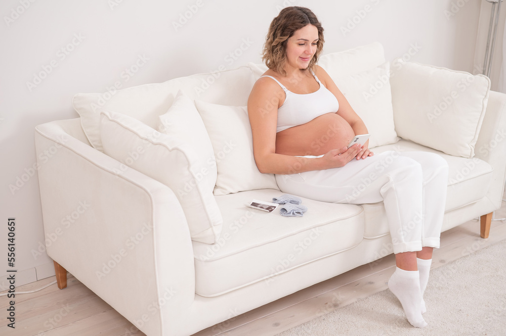 Pregnant woman sits on a white sofa and holds a smartphone. Fetal ultrasonic screening. Pregnant woman sits on a white sofa and holds a smartphone. Fetal ultrasonic screening.