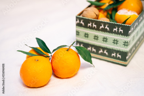 Several beautiful fresh tangerines with leaves are lying on a crumpled white bedspread. There is a box of tangerines and nuts nearby. New Year, atmosphere, comfort and warmth.