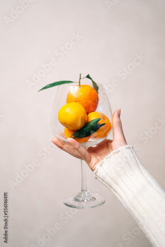 A beautiful female hand holds a glass glass, inside of which there are several tangerines with leaves on a light background. Vertical photo.