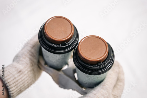 A woman in knitted beige mittens holds a cardboard stand with two cups of coffee on which there are chocolate cookies.