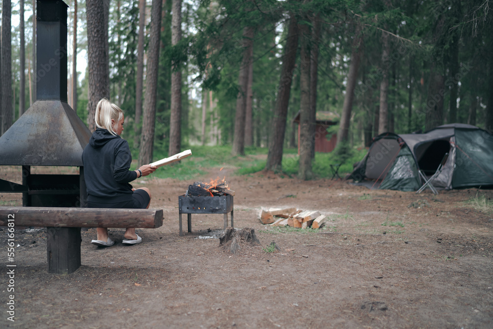 Camping in the forest. A girl throws firewood into a burning barbecue in nature, a tent is in the background. Camping in the forest. A girl throws firewood into a burning barbecue in nature, a tent is in the background.