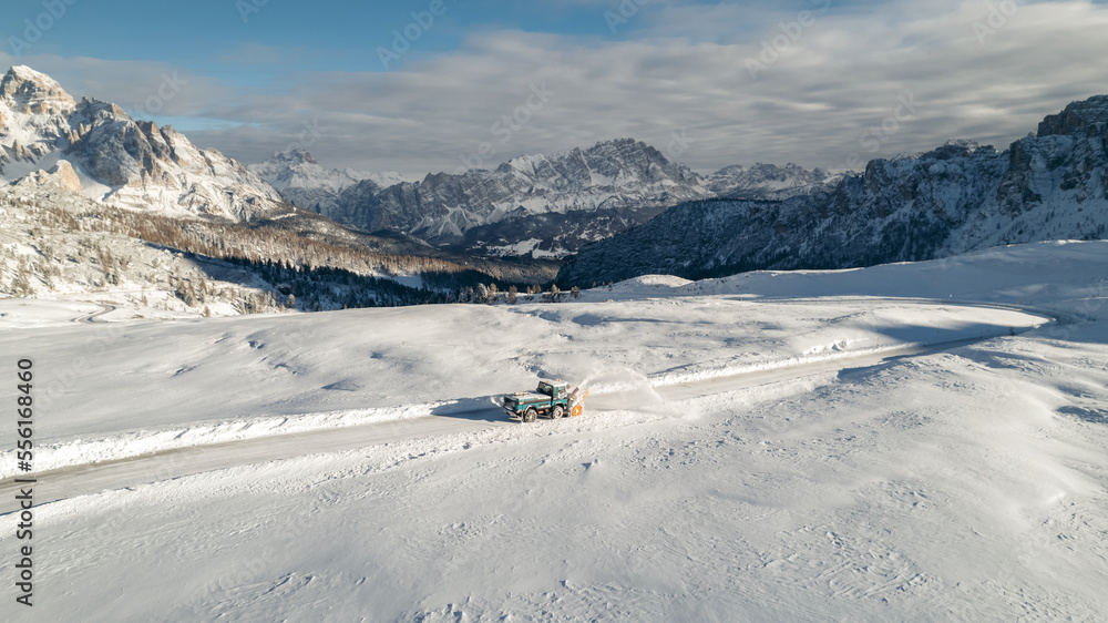 Snow Plow Truck cleaning and blowing the snow off the road in the province of Belluno in Italy. Breathtaking mountain view from drone. Cold sunny winter day.