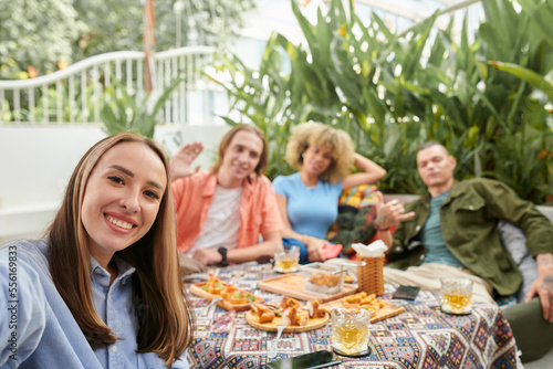 Cheerful young woman taking selfie with group of her friends when they are enjoying dinner in outdoor cafe