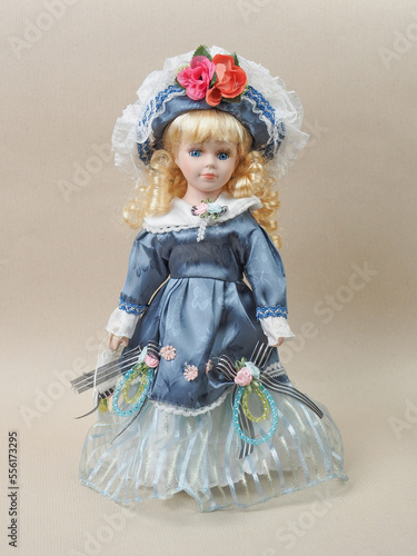 Vintage porcelain doll baby girl with blue eyes, blonde curls, wearing a blue hat decorated with red flowers and a blue satin dress.