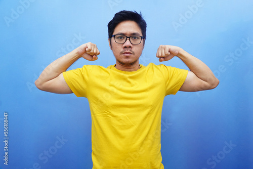 Excited Asian man showing strong gesture by raising his arms and muscles wearing yellow t-shirt and blue background. photo
