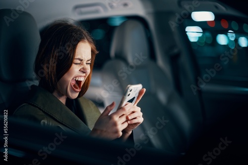 a close horizontal portrait of a stylish, luxurious woman in a leather coat sitting in a black car at night in the passenger seat, emotionally screaming while holding her smartphone © SHOTPRIME STUDIO