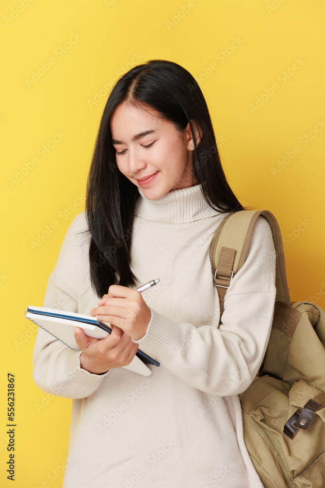 happy asian girl student with backpack writing in a notepad while standing with books isolated over yellow background happy asian girl student with backpack writing in a notepad while standing with books isolated over yellow background