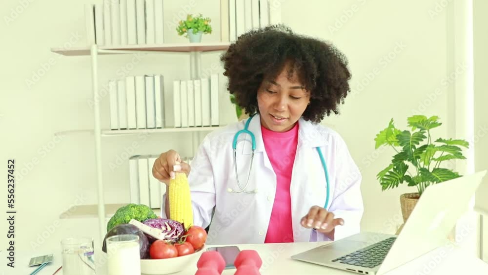 Cute african american female nutritionist sitting on her desk smiling and presenting organic vegetable corn online to patient to consume on laptop computer working in hospital office. Cute african american female nutritionist sitting on her desk smiling and presenting organic vegetable corn online to patient to consume on laptop computer working in hospital office.