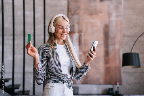 Beautiful blonde young Swedish woman in headphones broad smiles wide openes mouth and eyes holds credit card and phone amazed by discount, sale, Black Friday. Excited girl online purchase. photo