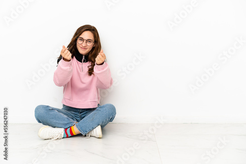 Young caucasian woman sitting on the floor isolated on white background making money gesture