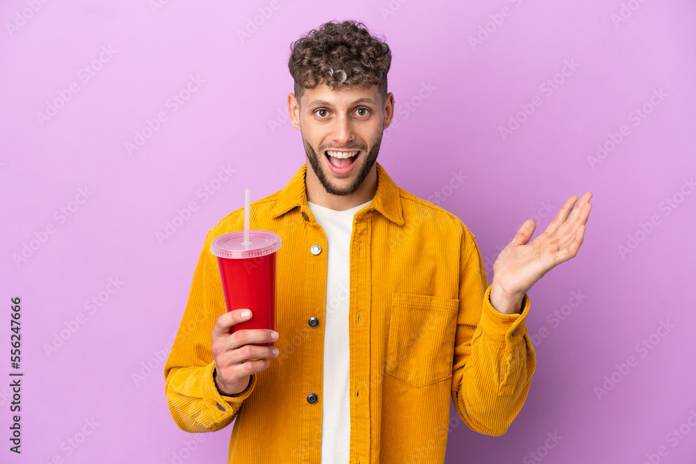 Young blonde man holding soda isolated on purple background with shocked facial expression Young blonde man holding soda isolated on purple background with shocked facial expression