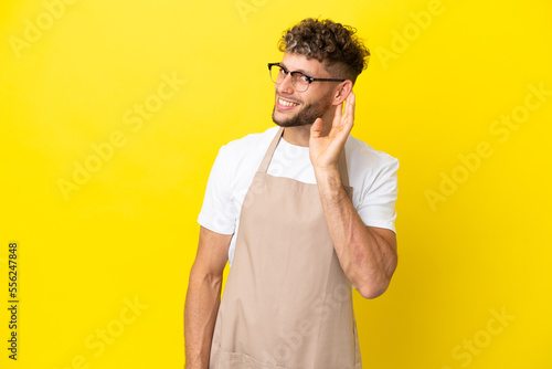 Restaurant waiter blonde man isolated on yellow background listening to something by putting hand on the ear