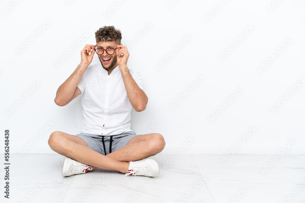 Young blonde man sitting on the floor isolated on white background with glasses and surprised Young blonde man sitting on the floor isolated on white background with glasses and surprised