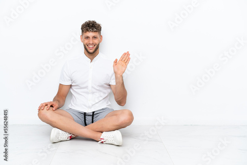 Young blonde man sitting on the floor isolated on white background saluting with hand with happy expression