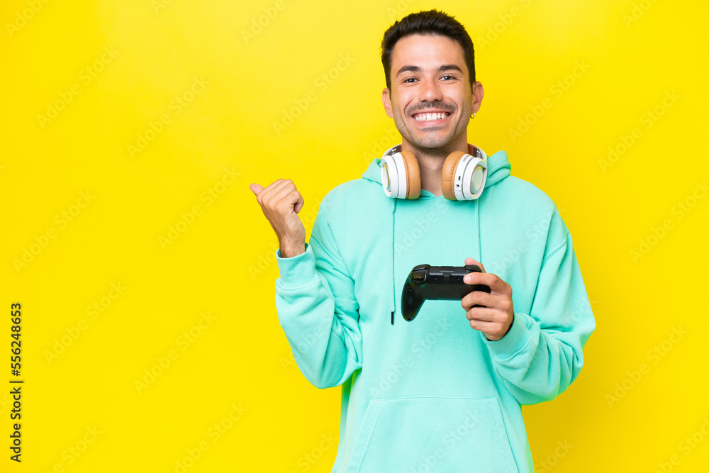 Young handsome man playing with a video game controller over isolated wall pointing to the side to present a product Young handsome man playing with a video game controller over isolated wall pointing to the side to present a product