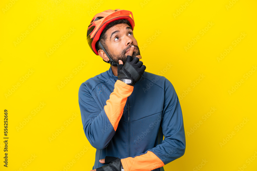 Fototapeta premium Young cyclist Brazilian man isolated on yellow background having doubts while looking up