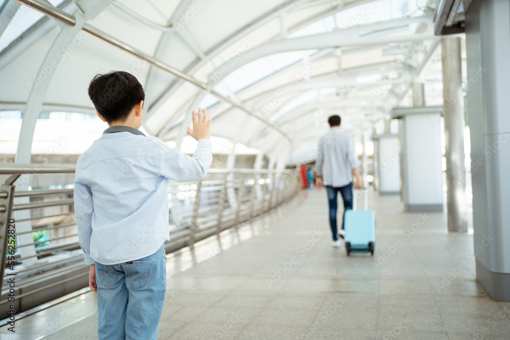 Asian little boy waves a hand and says goodbye to his father at the airport or railway terminal before father leaving.